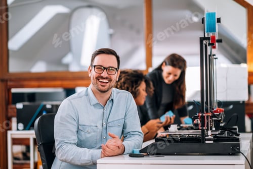 Preview: Portrait of a happy engineer with 3D printer, looking at camera.