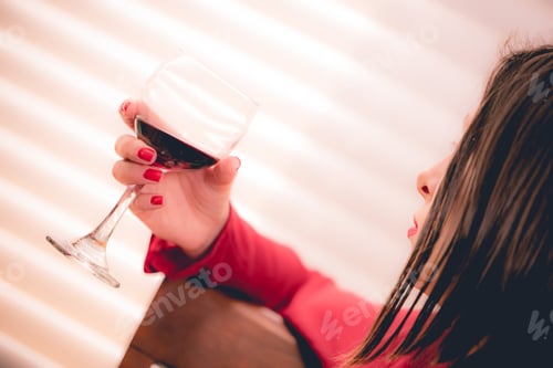 Preview: young millennial woman in red shirt and red fingernail polish enjoying a glass of red wine at home