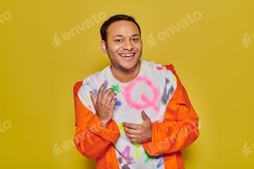Preview: gleeful indian man in orange jacket and diy t-shirt smiling and looking at camera on yellow backdrop