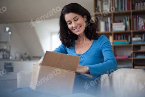 Preview: Smiling woman at home sitting on couch unpacking parcel