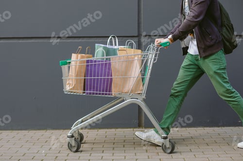 Preview: A man in bright trousers rolling a shopping cart.