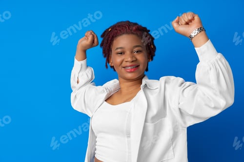 Preview: Portrait of a cheerful young african woman with hands raised against blue background