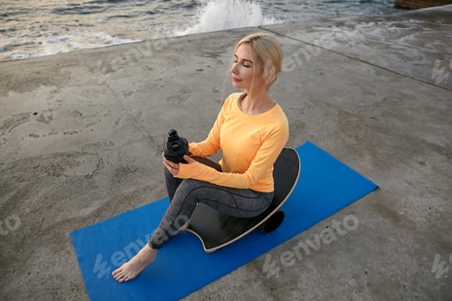 Preview: Young attractive sporty female with blonde hair making sports over seaside sitting on balance board