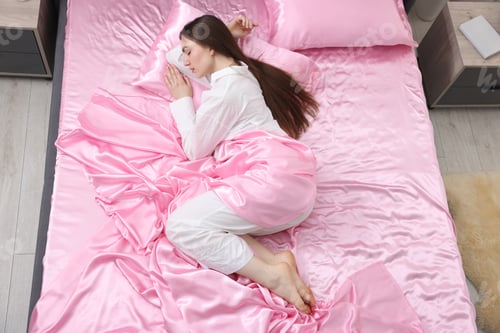 Preview: Woman sleeping in bed with pink silk linens indoors, top view