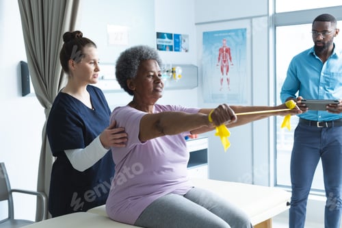 Preview: Diverse senior female patient exercising with band and female doctor advising in hospital room