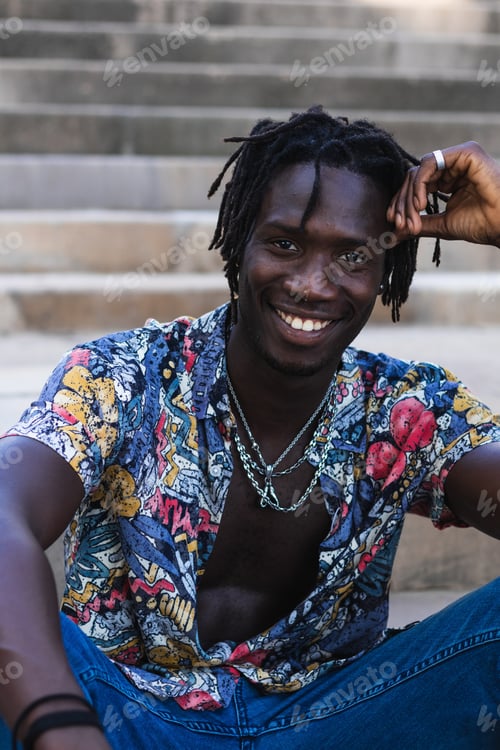 Preview: Cheerful African American male in colorful shirt and jeans sitting on stone steps and looking at