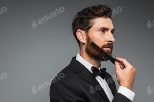 Preview: man in tuxedo brushing beard with comb and looking away isolated on grey