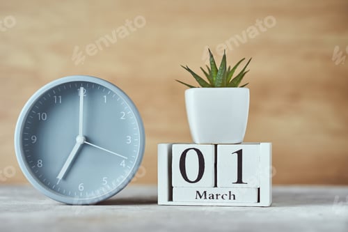 Preview: Alarm clock with wooden block calendar date 1st march and plant on the table