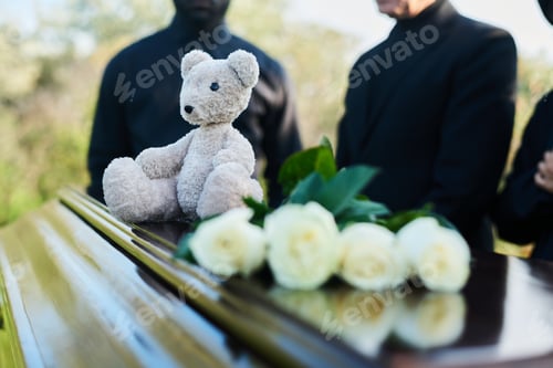 Preview: Focus on teddybear sitting on closed lid of coffin with bunch of white roses