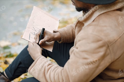 Preview: Black man sitting on a road and writing a sign for autostop