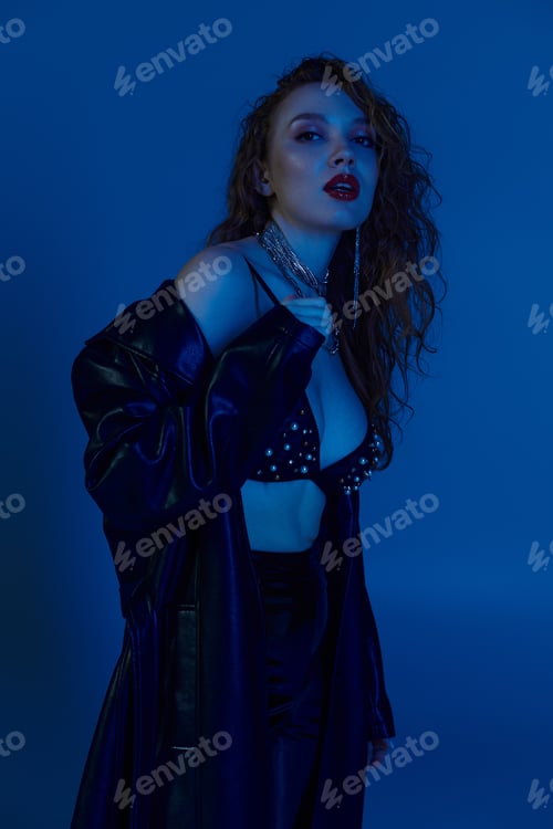 Preview: A stunning red-haired woman in stylish attire posing under vibrant blue lighting