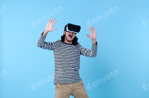 Preview: Excited Asian man wearing virtual reality goggles watching movies on blue background.