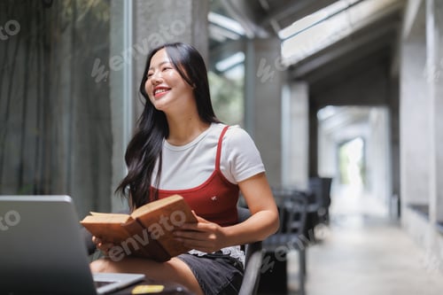 Preview: University student smiling and reading a book while studying with laptop