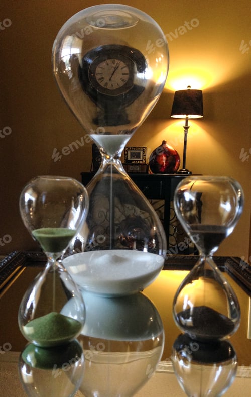 Preview: Hour glass reflected in mirror with wall clock in background and lamp making hourglass shape sand
