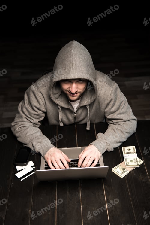 Preview: Young hacker in hoodie sitting at table and using laptop
