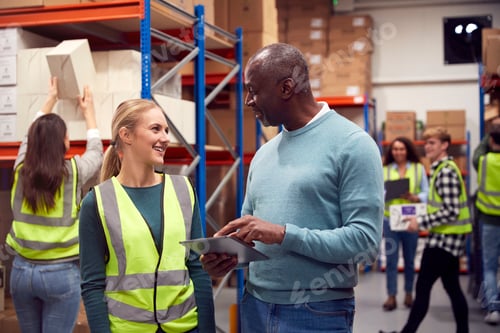 Preview: Female Intern With Team Leader Looking At Digital Tablet Inside Busy Warehouse Facility