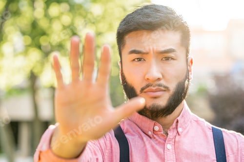 Preview: Sun illuminating an asian man in casual clothes gesturing stop with outstretched hand in a park