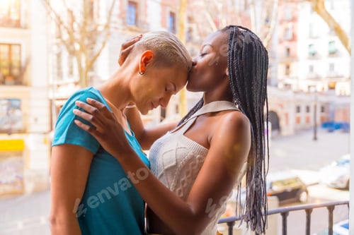 Preview: Beautiful diverse lesbian couple kissing on the forehead on the balcony at home, LGBT concept
