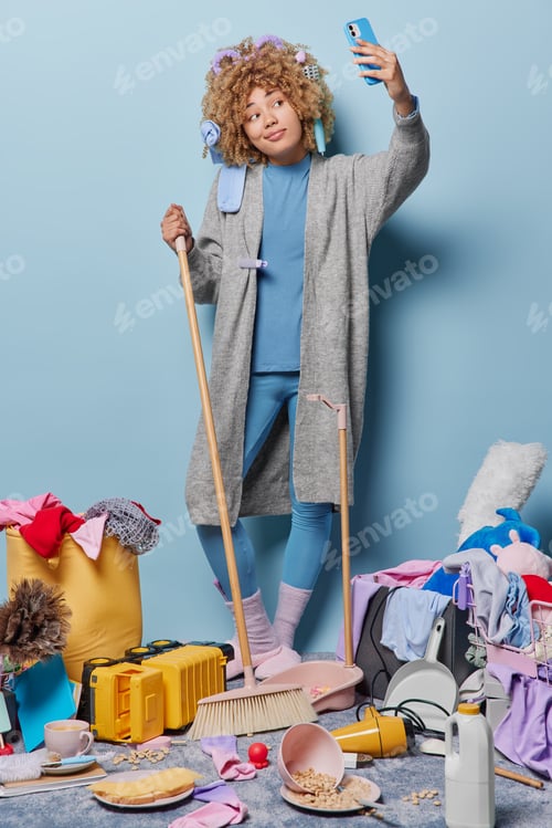 Preview: Vertical shot of curly haired housewife dressed in domestic clothes sweeps floor takes selfie via