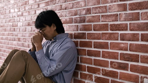 Preview: Christian Asian man is sitting against a wall, feeling sad, holding a cross and praying to God.