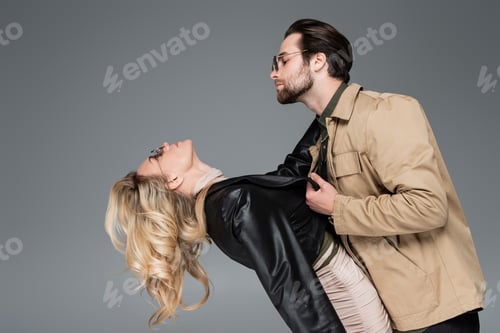 Preview: side view of stylish man in sunglasses pulling leather jacket of blonde woman isolated on grey
