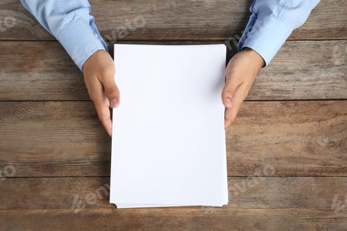 Preview: Man holding blank paper sheets for brochure at wooden table, top view. Mock up
