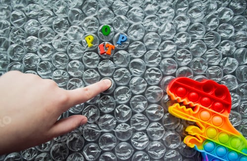 Preview: Toy pop it dinosaur on bubble wrap background with letters Pop it. Finger pushes bubble, dino looks
