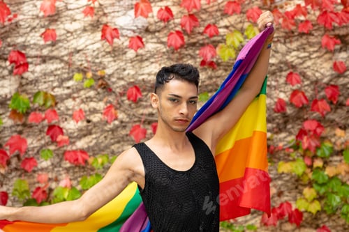 Preview: Portrait of biracial transgender man holding rainbow flag, with rocks and leaves in background