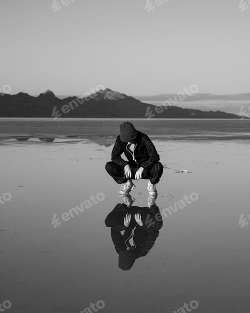 Preview: Person squatting by reflective water with mountains in background.
