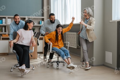 Preview: Happy office workers having fun riding on chairs in modern office