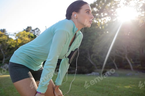 Preview: Exhausted female jogger listening to music and leaning on knees while exercising