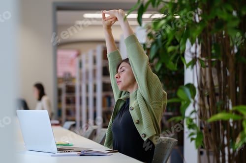 Preview: Tired female freelancer stretching arms relaxing while working on laptop in cozy coworking space