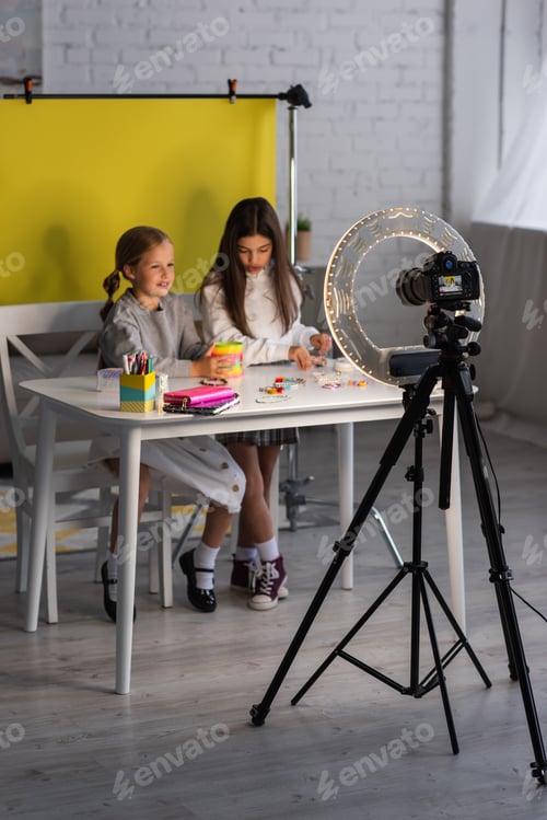 Preview: circle lamp and digital camera on tripod near preteen girls recording video blog on yellow