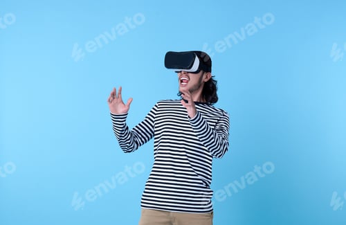 Preview: Excited Asian man wearing virtual reality goggles watching movies on blue background.