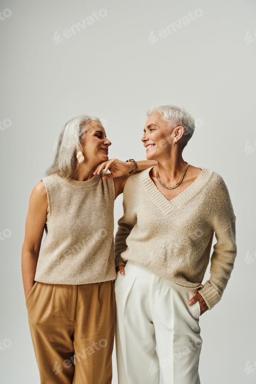 Preview: trendy senior women in pastel clothes looking at each other on grey, fashionable long-life friends