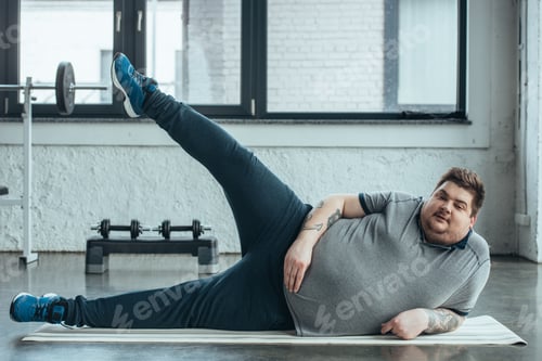 Preview: overweight tattooed man lying on fitness mat and stretching legs at gym
