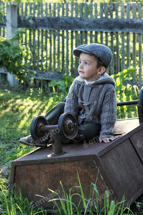 Preview: A cute 3-year-old boy is sitting in the village by the fence on an iron wheelbarrow