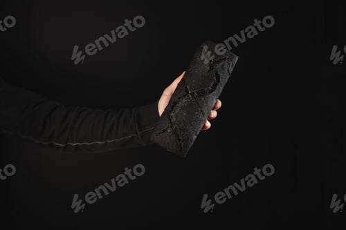 Preview: Man hand in black sweatshot holds homemade luxury rustic charcoal bread, isolated on black