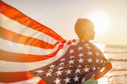 Preview: Back view candid authentic gen Z boy wrapped American flag looks at the sea on celebrate July 4th
