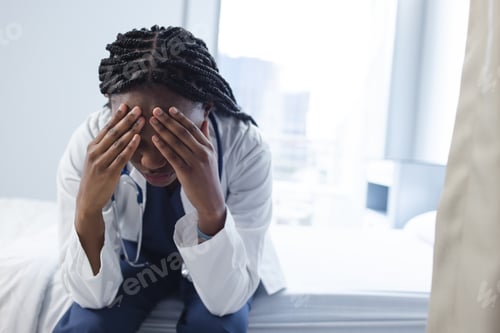 Preview: Tired african american female doctor wearing lab coat sitting on bed in hospital room, copy space