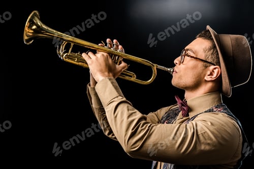 Preview: side view of young male jazzman in hat and eyeglasses playing on trumpet isolated on black