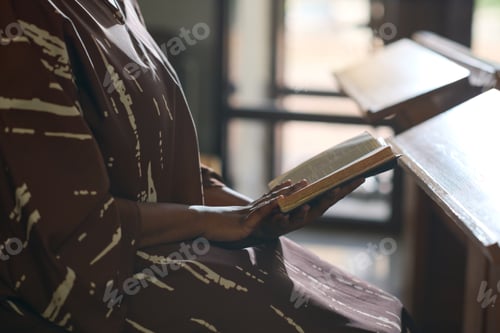 Preview: Side view of young black woman in casual dress sitting in church with Bible