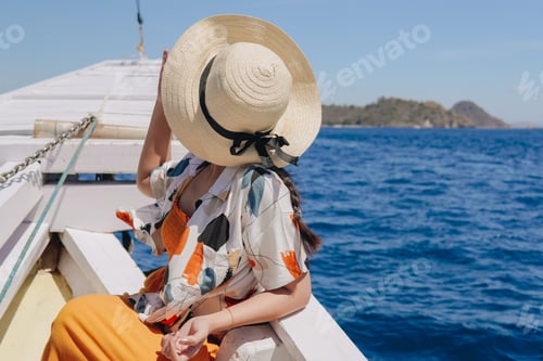 Vista previa: Vista posterior de una joven asiática con un sombrero de paja disfrutando del paisaje de Labuan Bajo en un barco.