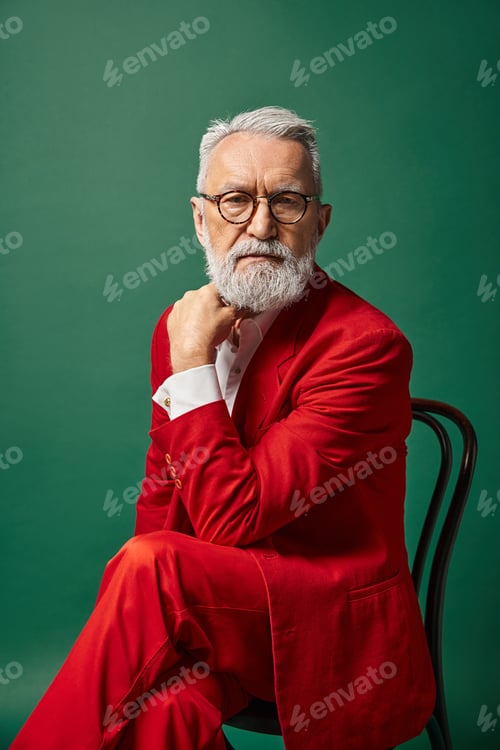 Preview: elegant handsome Santa in red suit posing on chair with his hand near face, Christmas concept