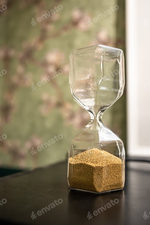 Preview: Close up of glass hourglass on blurred background.