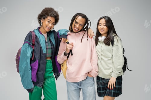 Preview: pretty multicultural women posing near trendy man with dreadlocks and backpack isolated on grey