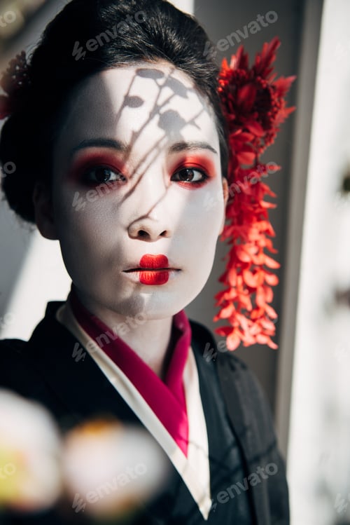 Preview: selective focus of sakura branches and beautiful geisha with red and white makeup in sunlight
