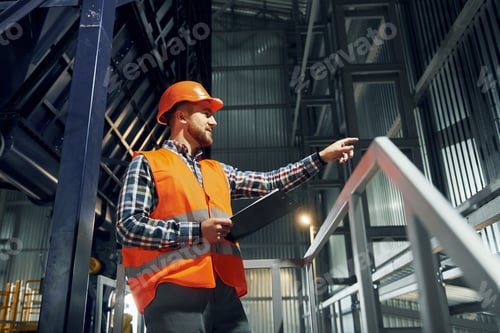 Preview: In orange colored hard hat. Construction worker in uniform is in the factory