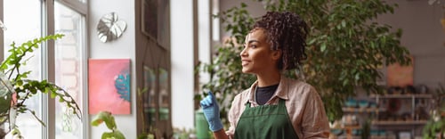 Preview: Attractive smiling florist stands near windows on background of her own floral decor studio