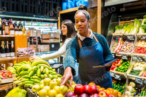 Preview: Saleswoman arranging fresh fruit in organic supermarket with customer choosing bananas